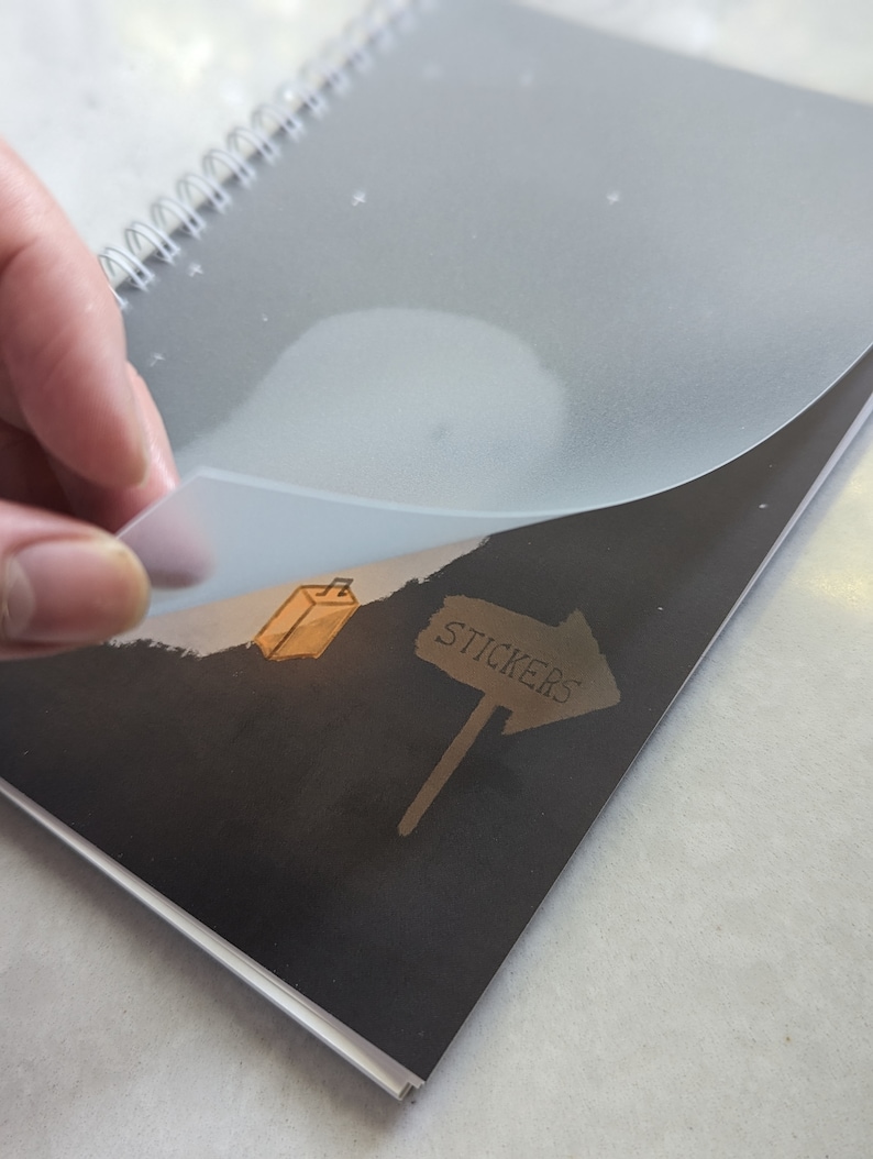 May include: A black spiral bound notebook with a clear plastic cover. The cover has a white cloud and stars design. The notebook has a black and white illustration of a sign that says "Stickers" with an arrow pointing to the right. The illustration also includes a small brown box.