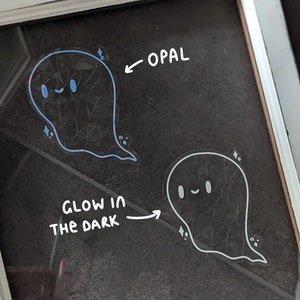 May include: Two ghost stickers, one blue and one white, on a black surface. The blue ghost sticker is labeled "OPAL" and the white ghost sticker is labeled "GLOW IN THE DARK".