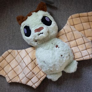 May include: A plush green bat toy with brown ears and large eyes. The bat has a waffle-like texture on its wings.