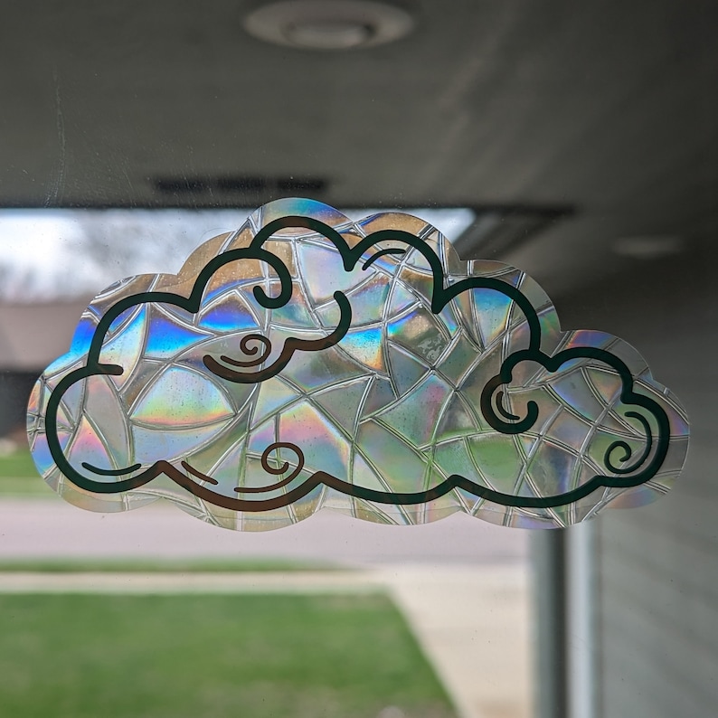 May include: A holographic sticker of a cloud with a rainbow iridescent finish. The cloud is outlined in black and has a whimsical design.