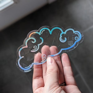 May include: A clear iridescent sticker with a blue and pink outline of a cloud with swirls. The sticker is designed to look like a stained glass window.