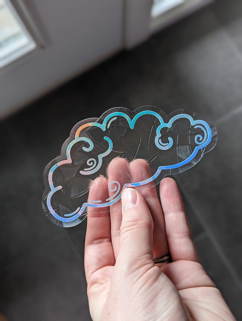 May include: A clear iridescent sticker with a blue and pink outline of a cloud with swirls. The sticker is designed to look like a stained glass window.