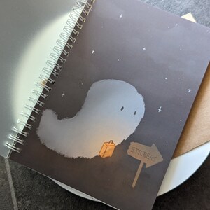 May include: A black spiral bound notebook with a ghost illustration on the cover. The ghost is white and has a small orange box next to it. A brown sign with the word "STICKERS" is pointing to the right.