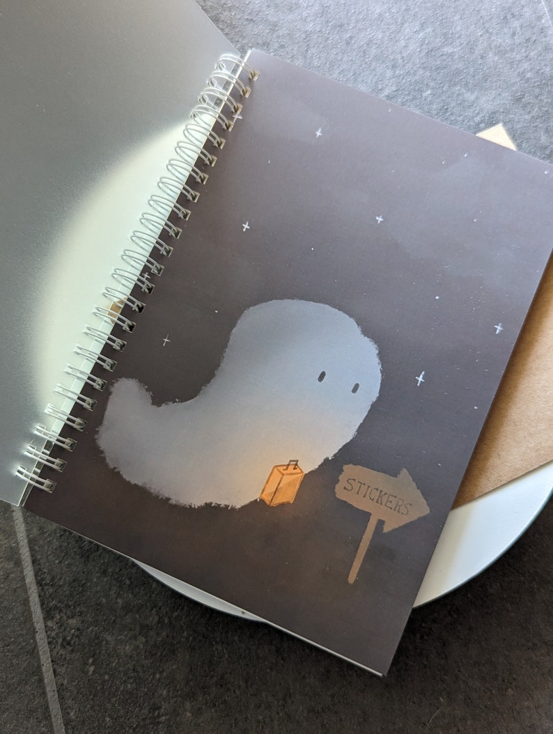 May include: A black spiral bound notebook with a ghost illustration on the cover. The ghost is white and has a small orange box next to it. A brown sign with the word "STICKERS" is pointing to the right.