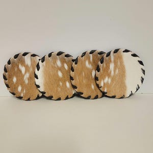 May include: Four round coasters made of brown and white spotted animal hide with black stitching around the edges.