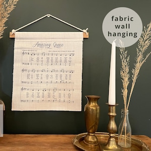 May include: A beige fabric wall hanging with the lyrics to the song "Amazing Grace" printed on it. The hanging is displayed on a dark green wall with a wooden dowel rod at the top.