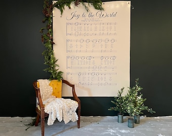 May include: A vintage wooden chair with a yellow cushion and a white lace doily. A large sheet of paper with the words "Joy to the World" and musical notes hangs on the wall. Small Christmas trees and greenery decorate the scene.