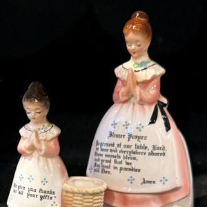 Puede incluir: Dos figuras de cerámica de mujeres en oración, una grande y otra pequeña, con una cesta de mimbre. Las figuras visten vestidos rosas y blancos. La figura más grande tiene el texto "Dinner Prayer".