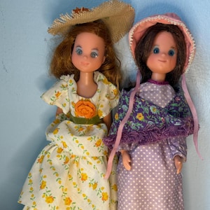 May include: Two vintage dolls are displayed, each wearing a bonnet or hat and a dress. One doll has red hair and a straw hat, wearing a white dress with yellow floral patterns. The other doll has brown hair and a pink-trimmed bonnet, wearing a purple dress.