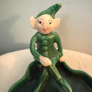 May include: A ceramic figurine of a seated elf with a green outfit and hat, and a white face with painted features. The elf is perched on a large, green leaf-shaped dish. The figurine has a vintage, handcrafted appearance.