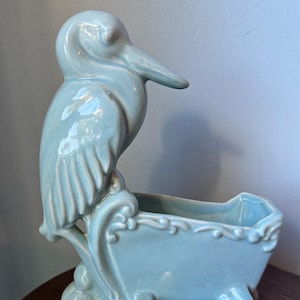 May include: A light blue ceramic planter shaped like a bird carrying a wheelbarrow. The planter has a glazed finish and intricate details.