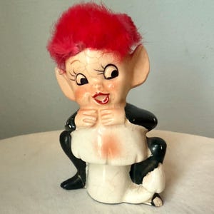 May include: A vintage ceramic figurine of a seated elf with a red, fluffy hairpiece. The elf has large ears, a smiling face, and is wearing a black and white outfit. The figurine is perched on a white mushroom.