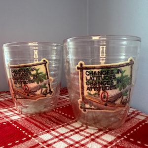 Pode incluir: Dois copos de plástico transparente com um patch decorativo com uma rede, uma palmeira e o texto "Changes in Latitudes, Changes in Attitudes". Os copos estão sobre uma superfície xadrez vermelha e branca.