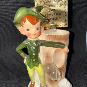 Puede incluir: Una figura de duende vintage con sombrero y atuendo verdes, pantalones amarillos y zapatos rojos. La figura sostiene un letrero que dice "Helpful Kitchen Pixie Says, USE THIS CLIP for RECIPE CARDS MESSAGES."