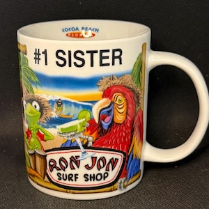 K&ouml;nnte beinhalten: Ein wei&szlig;er Keramikbecher mit dem Text #1 SISTER und COCOA BEACH FLORIDA. Die Grafik zeigt einen gr&uuml;nen Frosch, einen roten Papagei, einen Surfer auf einer Welle und ein Cocktailglas. Das Logo RON JON SURF SHOP ist unten in der Mitte aufgedruckt.