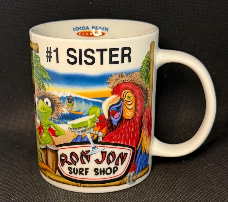 K&ouml;nnte beinhalten: Ein wei&szlig;er Keramikbecher mit dem Text #1 SISTER und COCOA BEACH FLORIDA. Die Grafik zeigt einen gr&uuml;nen Frosch, einen roten Papagei, einen Surfer auf einer Welle und ein Cocktailglas. Das Logo RON JON SURF SHOP ist unten in der Mitte aufgedruckt.