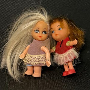 May include: Two vintage miniature dolls. One doll has long blonde hair, a purple dress with lace trim, and blue eyes. The other doll has short brown hair, a red dress with a white trim, and blue eyes. Both dolls are standing.