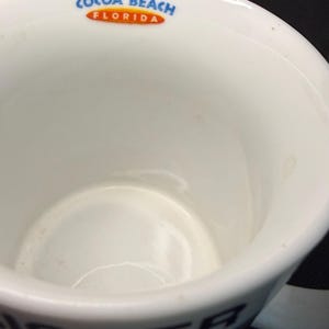 K&ouml;nnte beinhalten: Ein wei&szlig;er Keramikbecher mit dem Text COCOA BEACH FLORIDA in Blau und Orange am inneren Rand. Die Au&szlig;enseite des Bechers zeigt teilweise schwarze Buchstaben, die ISTER in der N&auml;he des Bodens auf wei&szlig;em Hintergrund bilden.