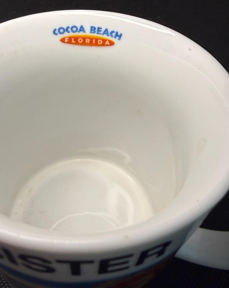 K&ouml;nnte beinhalten: Ein wei&szlig;er Keramikbecher mit dem Text COCOA BEACH FLORIDA in Blau und Orange am inneren Rand. Die Au&szlig;enseite des Bechers zeigt teilweise schwarze Buchstaben, die ISTER in der N&auml;he des Bodens auf wei&szlig;em Hintergrund bilden.