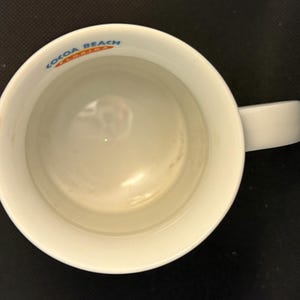 K&ouml;nnte beinhalten: Ein wei&szlig;er Keramikbecher, von direkt oben betrachtet, mit einem Henkel auf der rechten Seite. Der innere Rand zeigt den blau-orangen Schriftzug "COCOA BEACH FLORIDA", der entlang des Rands der kreisf&ouml;rmigen &Ouml;ffnung aufgedruckt ist.