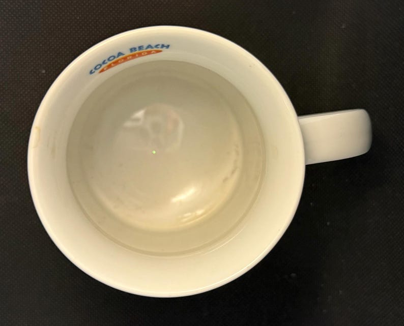 K&ouml;nnte beinhalten: Ein wei&szlig;er Keramikbecher, von direkt oben betrachtet, mit einem Henkel auf der rechten Seite. Der innere Rand zeigt den blau-orangen Schriftzug "COCOA BEACH FLORIDA", der entlang des Rands der kreisf&ouml;rmigen &Ouml;ffnung aufgedruckt ist.