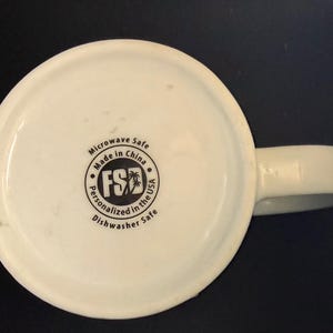K&ouml;nnte beinhalten: Die Unterseite einer wei&szlig;en Keramiktasse mit einem kreisf&ouml;rmigen schwarzen Logo, das den Text FSD und eine Palme zeigt. Der Text lautet Microwave Safe, Made in China, Personalized in the USA und Dishwasher Safe um das Logo herum.