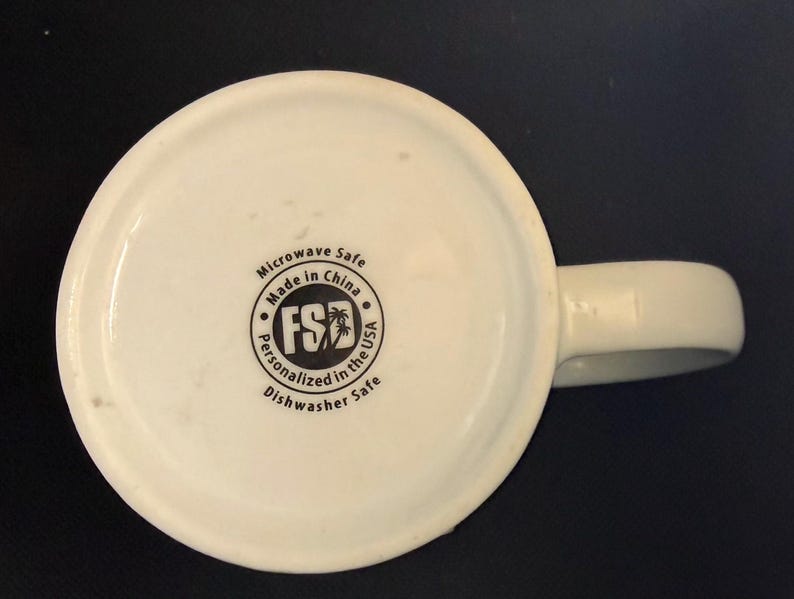 K&ouml;nnte beinhalten: Die Unterseite einer wei&szlig;en Keramiktasse mit einem kreisf&ouml;rmigen schwarzen Logo, das den Text FSD und eine Palme zeigt. Der Text lautet Microwave Safe, Made in China, Personalized in the USA und Dishwasher Safe um das Logo herum.