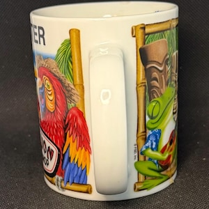 K&ouml;nnte beinhalten: Ein wei&szlig;er Keramikbecher mit einer bunten Grafik eines roten Papageis mit Strohhut und eines gr&uuml;nen Frosches, der eine kleine Gitarre spielt. Das Design zeigt bambusartige Rahmen, eine Tiki-Figur sowie den Text "TER" und "OON SHOP".
