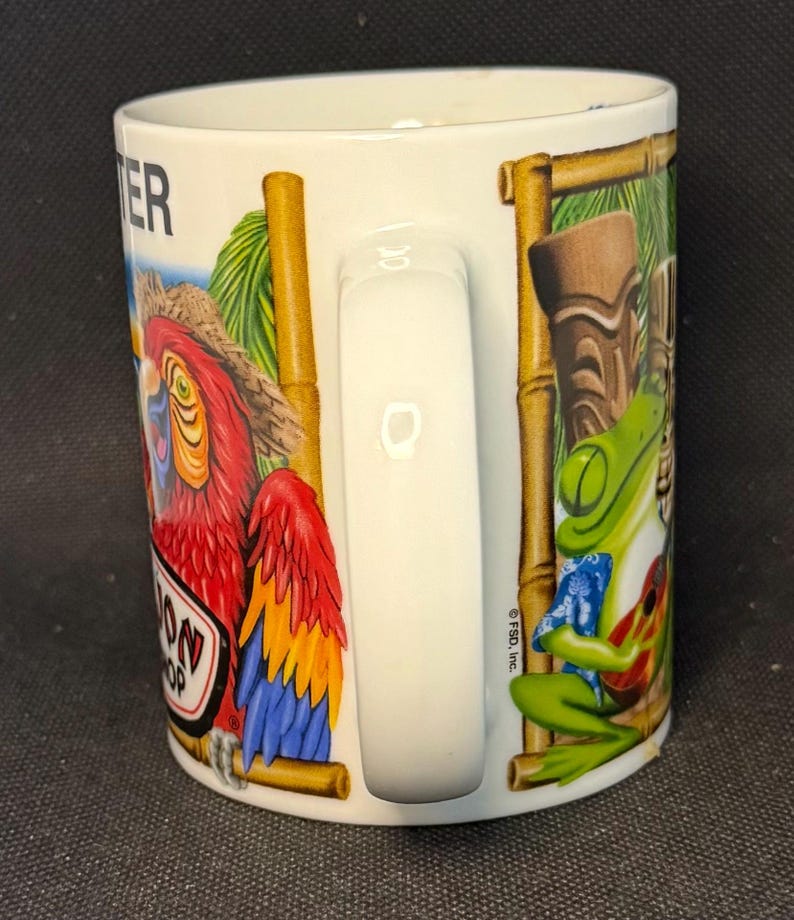 K&ouml;nnte beinhalten: Ein wei&szlig;er Keramikbecher mit einer bunten Grafik eines roten Papageis mit Strohhut und eines gr&uuml;nen Frosches, der eine kleine Gitarre spielt. Das Design zeigt bambusartige Rahmen, eine Tiki-Figur sowie den Text "TER" und "OON SHOP".