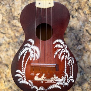 May include: A brown ukulele with a white painted tropical scene featuring palm trees and people in a boat. The ukulele has four strings and a sound hole. The instrument is made of wood.