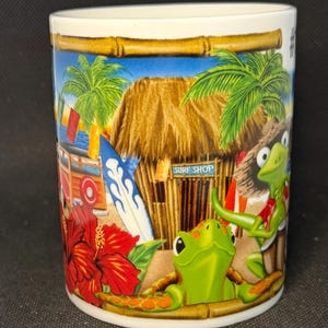 K&ouml;nnte beinhalten: Ein wei&szlig;er Keramikbecher mit einer farbenfrohen tropischen Szene, die einen Surfshop mit Strohdach, Palmen, ein blaues Surfbrett, eine rote Hibiskusbl&uuml;te und zwei gr&uuml;ne Schildkr&ouml;ten zeigt. Eine Schildkr&ouml;te tr&auml;gt einen Strohhut.
