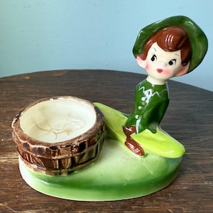 Puede incluir: Figura de cerámica vintage que representa un elfo con traje y sombrero verdes, con un recipiente en forma de barril marrón. La figura está sobre una base verde, mostrando un diseño caprichoso con un toque de nostalgia.