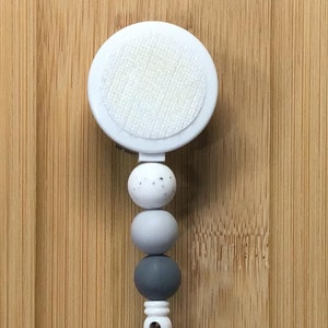 May include: A white retractable badge reel with a gray, white, and black beaded chain. The reel has a white circle with a gray and white speckled pattern.