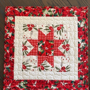 May include: A square quilted placemat with a Christmas theme. The placemat features a central star design in red gingham, surrounded by white squares with floral patterns and a red poinsettia border.
