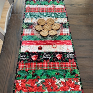 May include: A quilted Christmas table runner with alternating fabric strips. The runner features red, green, and white patterns with holiday motifs, including poinsettias, plaid, and the text "Happy Holidays" and "Peace". A clear glass plate holds several cookies.