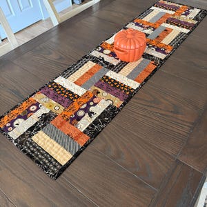 May include: A rectangular quilted table runner featuring a patchwork design of Halloween-themed fabrics in orange, black, purple, and cream, with a small orange ceramic pumpkin-shaped lidded container placed on top of the fabric surface.