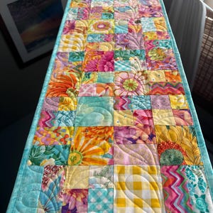 May include: A colorful, quilted table runner featuring a patchwork design. The runner showcases a variety of floral and geometric patterns in shades of yellow, orange, pink, and teal. The edges are finished with a solid teal border, adding a cohesive look.