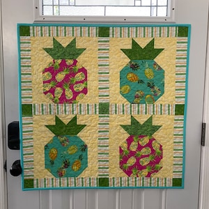 May include: A quilted wall hanging with four pineapple designs. The pineapples are made from fabric with a yellow, green, and pink pineapple pattern. The background fabric is yellow with white stripes. The wall hanging is framed with a turquoise border.