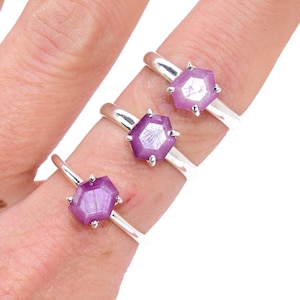 May include: Three silver rings each feature a hexagonal, faceted, light purple gemstone. The rings are displayed on a person's fingers against a white background. The gemstones have a reflective quality.