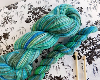 Könnte beinhalten: Ein Knäuel mehrfarbiger Wolle in Blau-, Grün- und Weißtönen, mit zwei Stricknadeln aus Holz. Die Wolle ist vor einem floral gemusterten Hintergrund drapiert, was auf ein Handwerks- oder Hobbythema hindeutet.