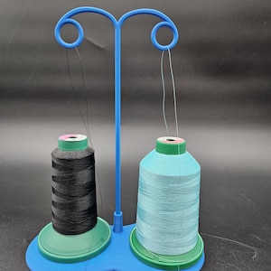 May include: A blue thread spool holder with two spools of thread. One spool is black, and the other is light blue. The spools sit on green bases, and the holder has a curved design with loops at the top to guide the thread.