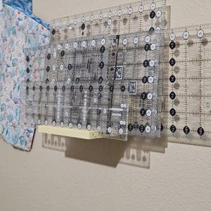 May include: A light yellow wall-mounted storage rack holding several clear acrylic quilting rulers. The rulers feature black grid lines, numerical markings, and circular patterns. A piece of fabric with a blue and white sea life print hangs nearby.