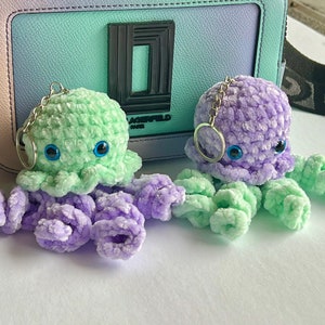 Puede incluir: Dos llaveros de pulpo de crochet con ojos azules. Un pulpo es verde y el otro es morado.