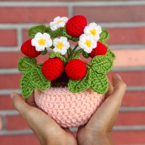 Op de afbeelding: Een gehaakte aardbeienplant in een roze pot. De plant heeft groene bladeren, rode aardbeien en witte bloemen met gele centra.