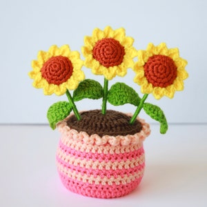 May include: A crocheted sunflower plant with three yellow flowers and green leaves in a pink and brown striped pot.
