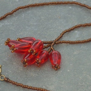 Peut inclure: Un collier de perles brun avec un pendentif en perles de verre rouge. Le pendentif est composé de nombreuses petites perles en forme de goutte.
