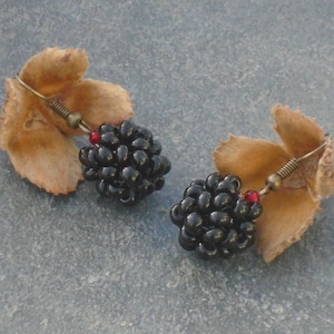 May include: A pair of earrings featuring black bead clusters that resemble blackberries, with a red bead accent on each earring. The earrings are displayed on a brown, textured surface.