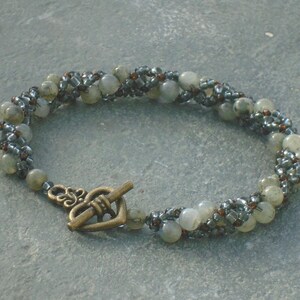 May include: A gray and silver beaded bracelet with a heart-shaped clasp. The bracelet is made with small, round beads and larger, oval-shaped beads.