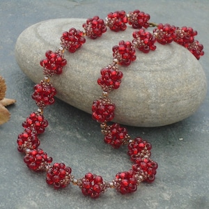 Peut inclure: Un collier composé de petites perles rouges rondes disposées en grappes. Les perles sont enfilées avec un fil métallique fin. Le collier est d'un rouge profond et est présenté sur une surface de pierre grise.