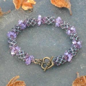 May include: A purple and gray beaded bracelet with a heart-shaped clasp. The bracelet is made with small, faceted beads and has a delicate, feminine look.